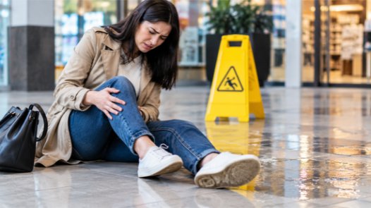 A woman sitting on a wet floor in a shopping mall holding her injured knee in pain next to a yellow wet floor caution sign, representing a slip and fall accident