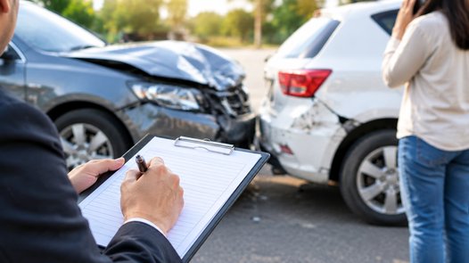 An insurance adjuster writing notes on a clipboard at the scene of a two-car accident, with a damaged sedan and SUV in the background, representing a fault assessment after a car accident